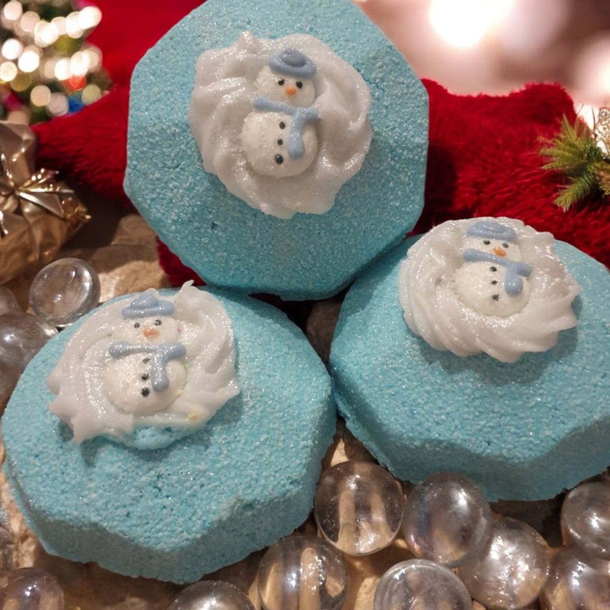 Three blue bath bombs with a white snowman design on top, displayed against a Christmas-themed background with red and green decorations.