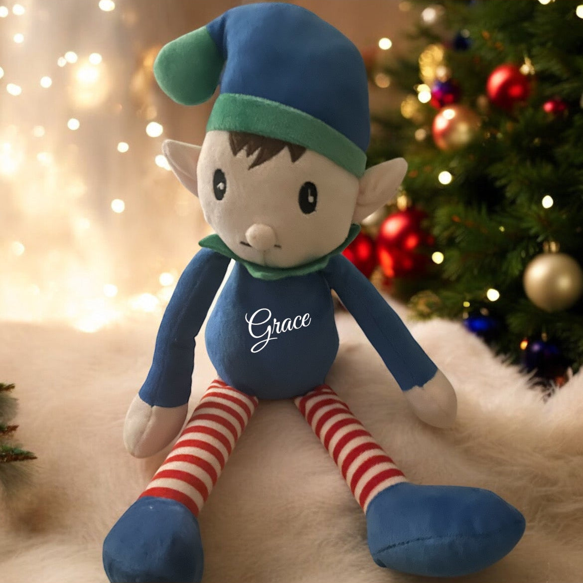 A blue personalised elf toy with a green hat and red and white striped legs, sitting on a white surface with a blurred Christmas tree background.