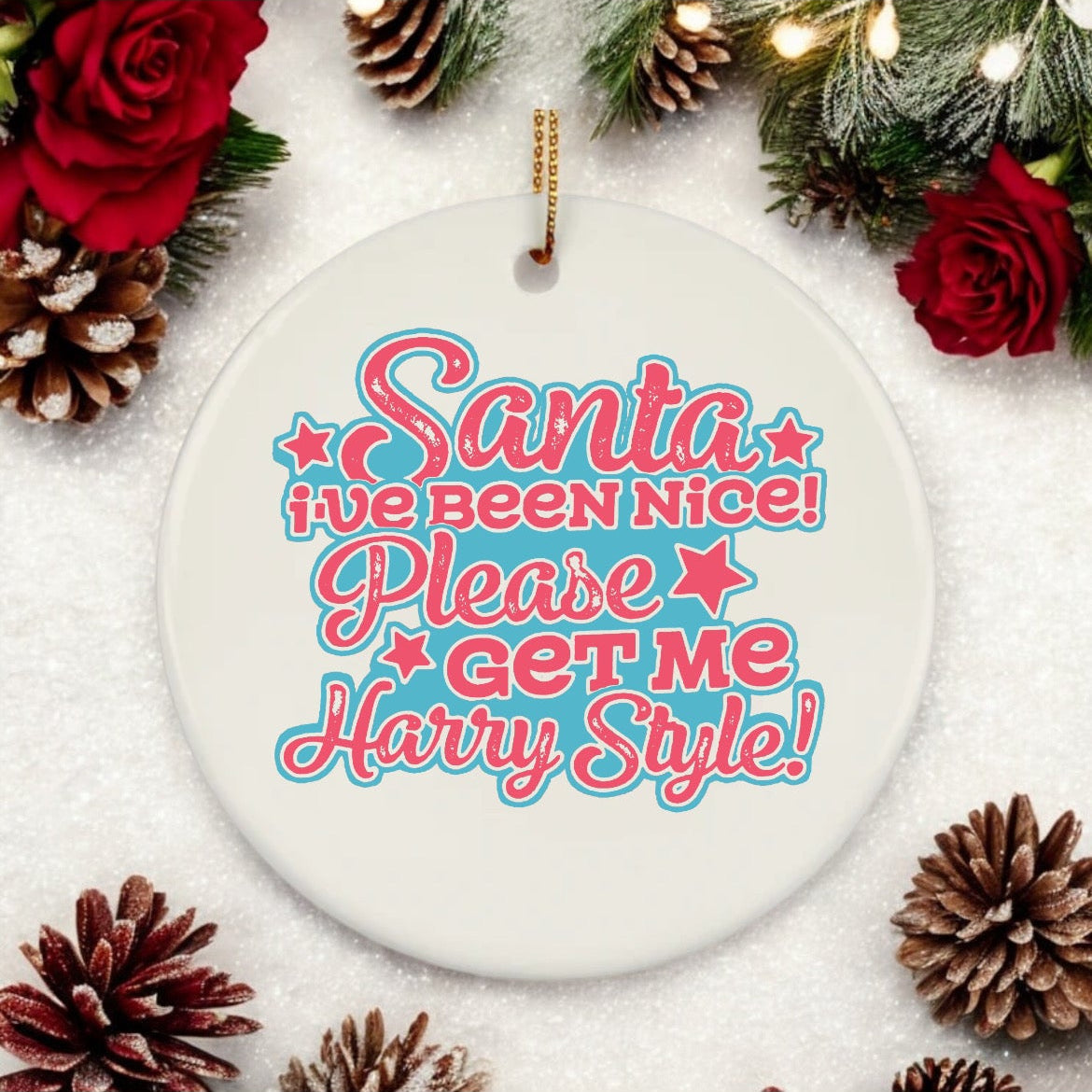 A ceramic bauble with text 'Santa's been nice! Please get me Harry Styles!' surrounded by Christmas decorations like pine cones and red and green lights.
