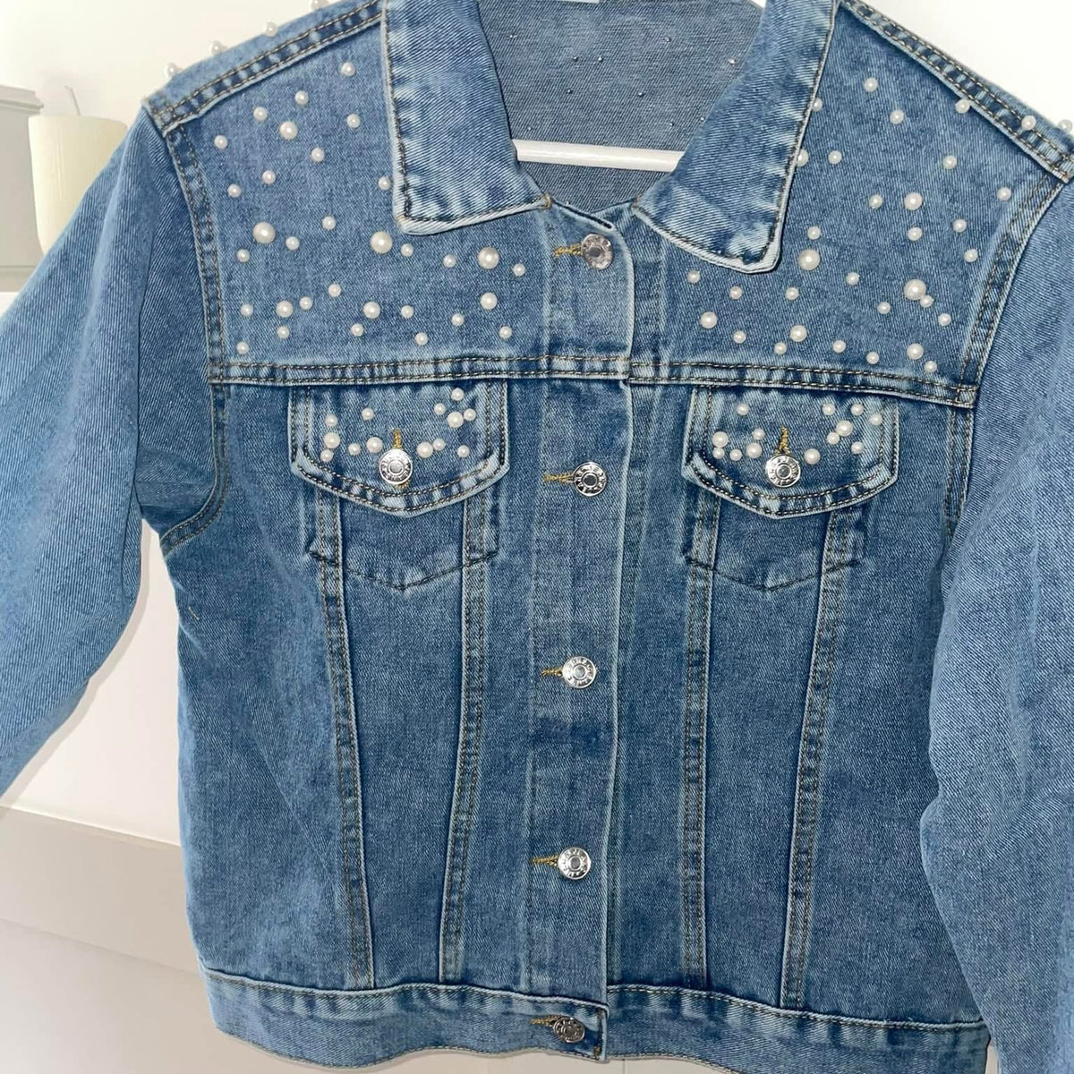 A denim jacket with pearl detail on the collar and chest area, displayed on a white hanger.