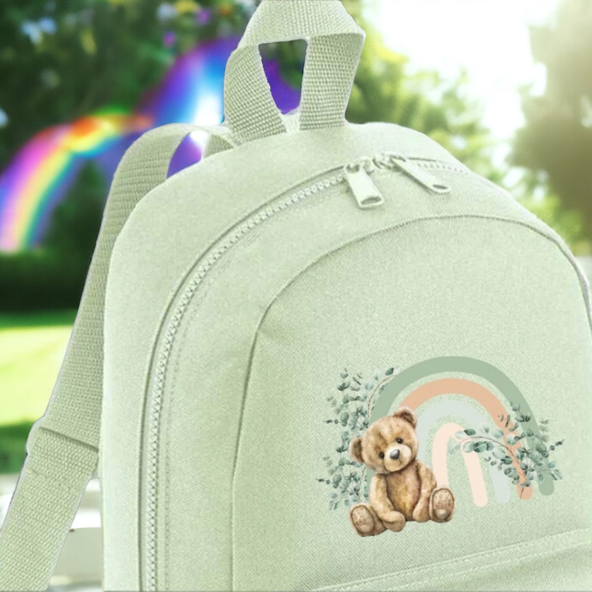 Mint green backpack featuring a teddy bear design with a rainbow motif, suitable for children.