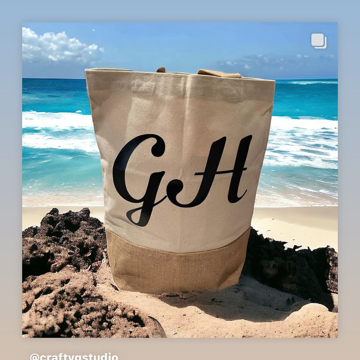 A beige cotton tote beach bag with a large 'G' in the center, placed on a rocky surface with a blue ocean background.