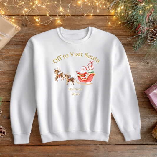 A white crewneck sweatshirt with a personalized Christmas graphic of Santa and reindeer, laid flat on a rustic wooden surface surrounded by holiday decorations like pine branches, pinecones, and wrapped gift boxes.