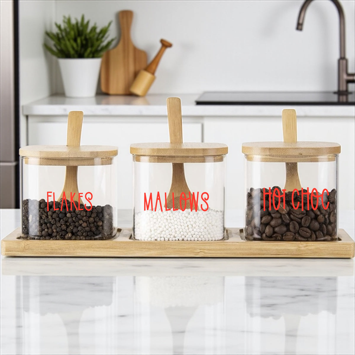 Three glass jars with wooden lids on a kitchen counter, labeled 'Flakes', 'Malows', and 'Hot Choc'.