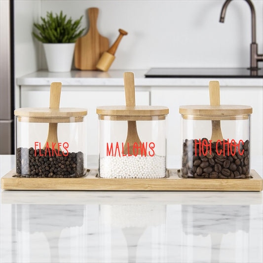 Three glass jars with wooden lids on a kitchen counter, labeled 'Flakes', 'Malows', and 'Hot Choc'.