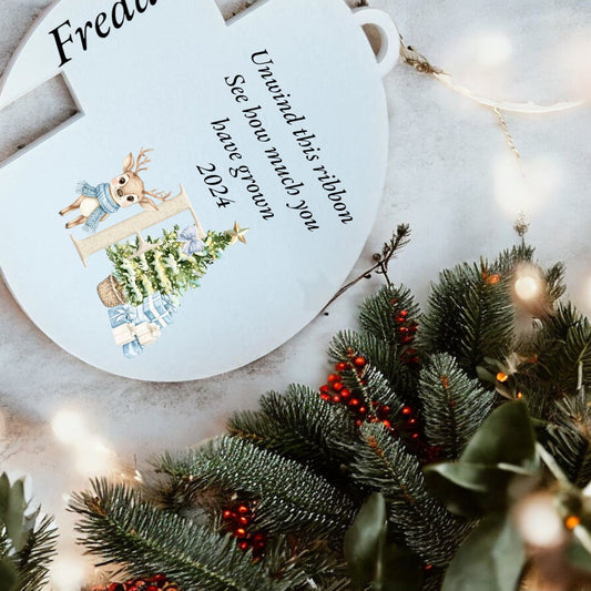 A personalized Christmas ornament with a reindeer, tree, and custom text in the center, surrounded by artificial holly and berries on a white background.
