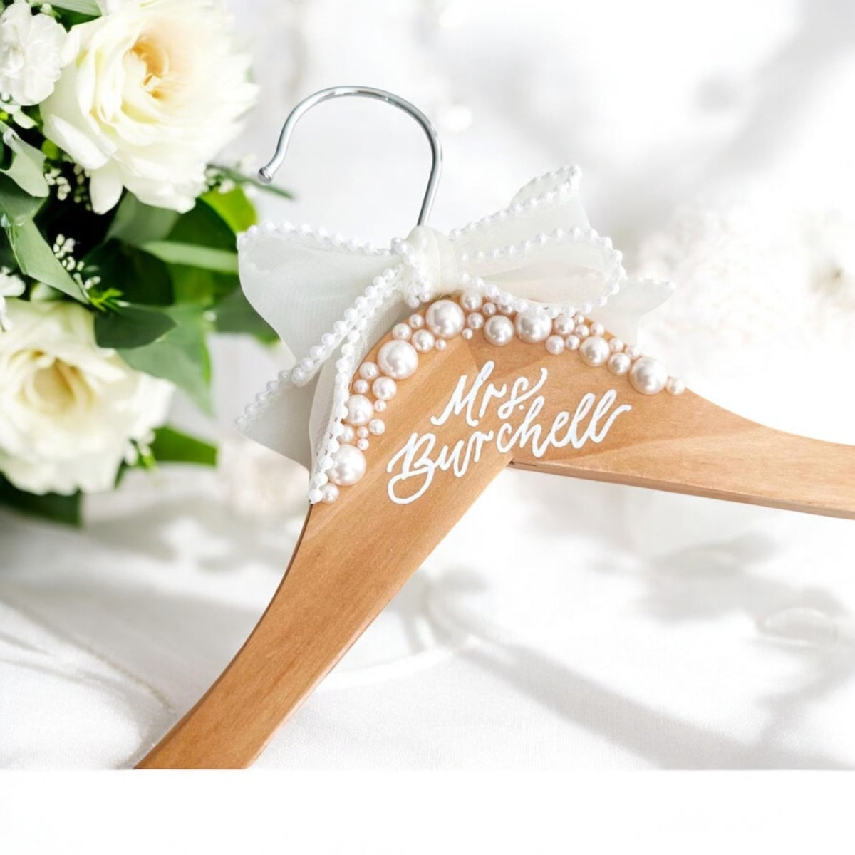 A wooden bridal hanger decorated with pearls and the name 'Mrs. Burwell' printed on it, displayed alongside a bouquet of flowers.