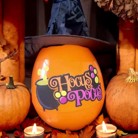 A personalized orange stuffable egg with 'BOO' text design, decorated with pumpkin and witch illustrations, displayed on a table with pumpkins and a witch's hat in the background.