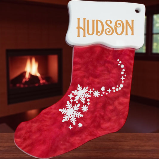Red Christmas stocking with white snowflake design and 'Hudson' name tag, placed in front of a fireplace.