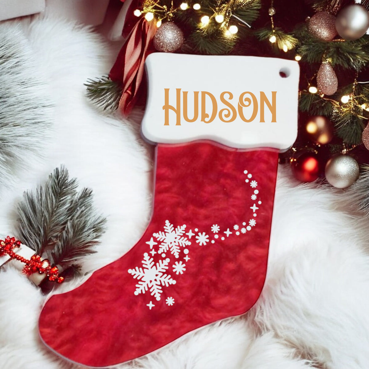 Red Christmas stocking with snowflake design and 'Hudson' name tag, placed under a decorated tree.