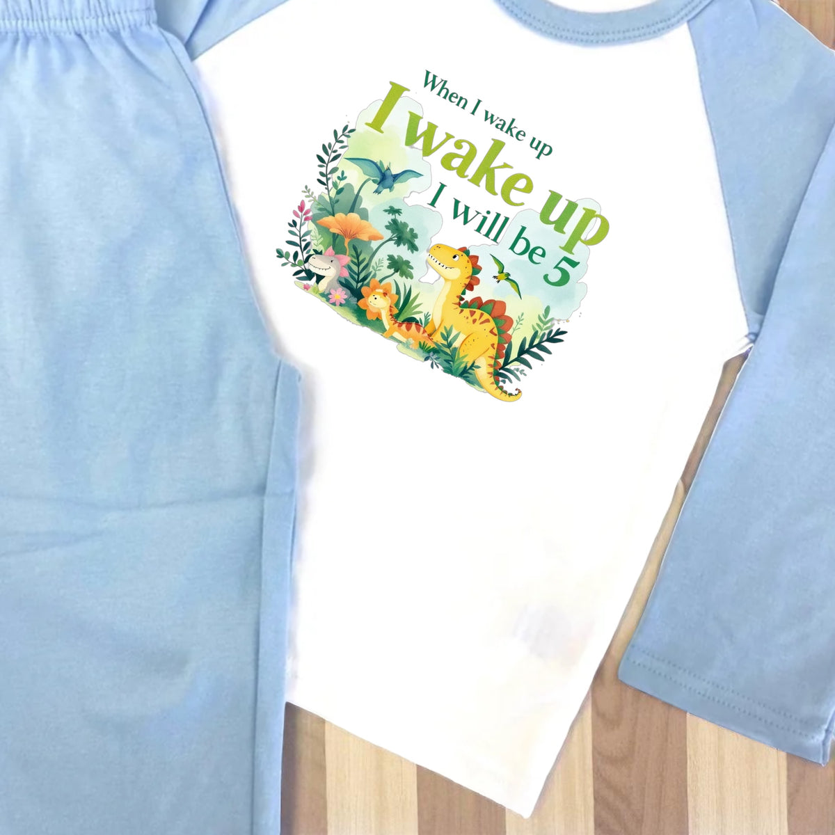 Children's outfit with a dinosaur-themed shirt and light blue pants on a wooden surface.