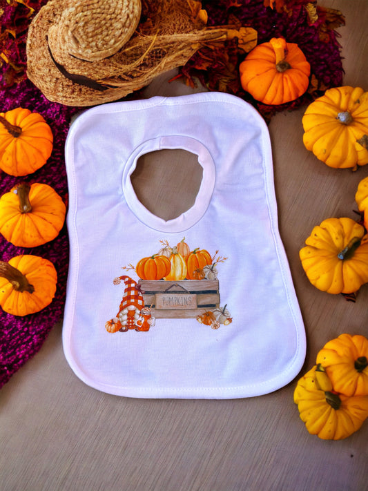 A white bib with a watercolor-like pumpkin patch design, featuring colors such as orange and yellow, presented on a table with pumpkins around it.
