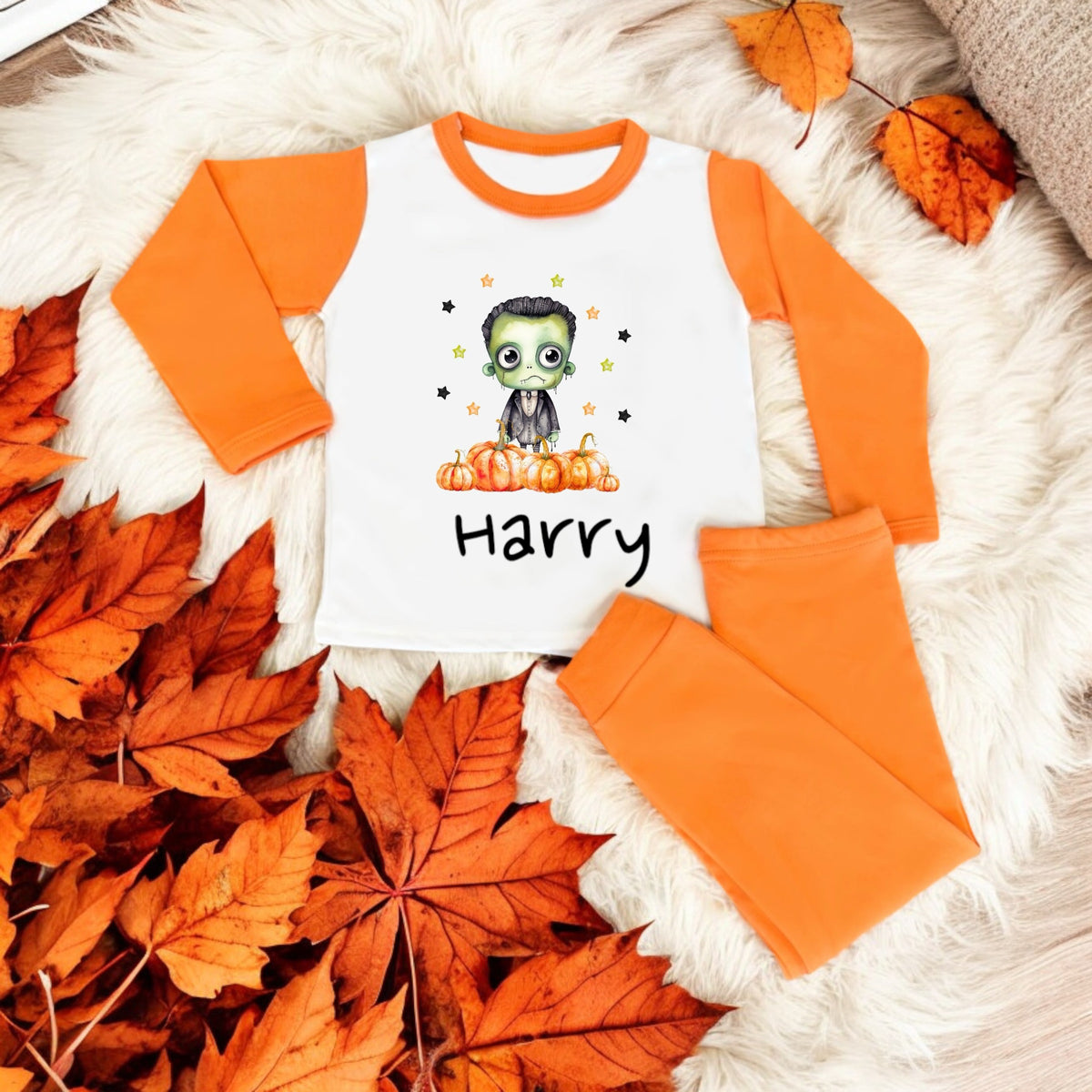 A set of children's pajamas with a Halloween monster design, laid out on a furry surface with autumn leaves scattered around. The pajamas are orange with a character illustration and the name 'Harry' printed on the front.