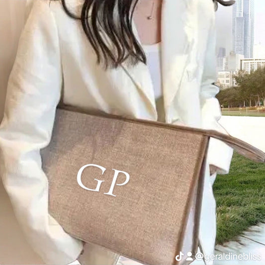 A person wearing a white jacket is holding a beige personalized clutch bag with the letter 'G' on it.