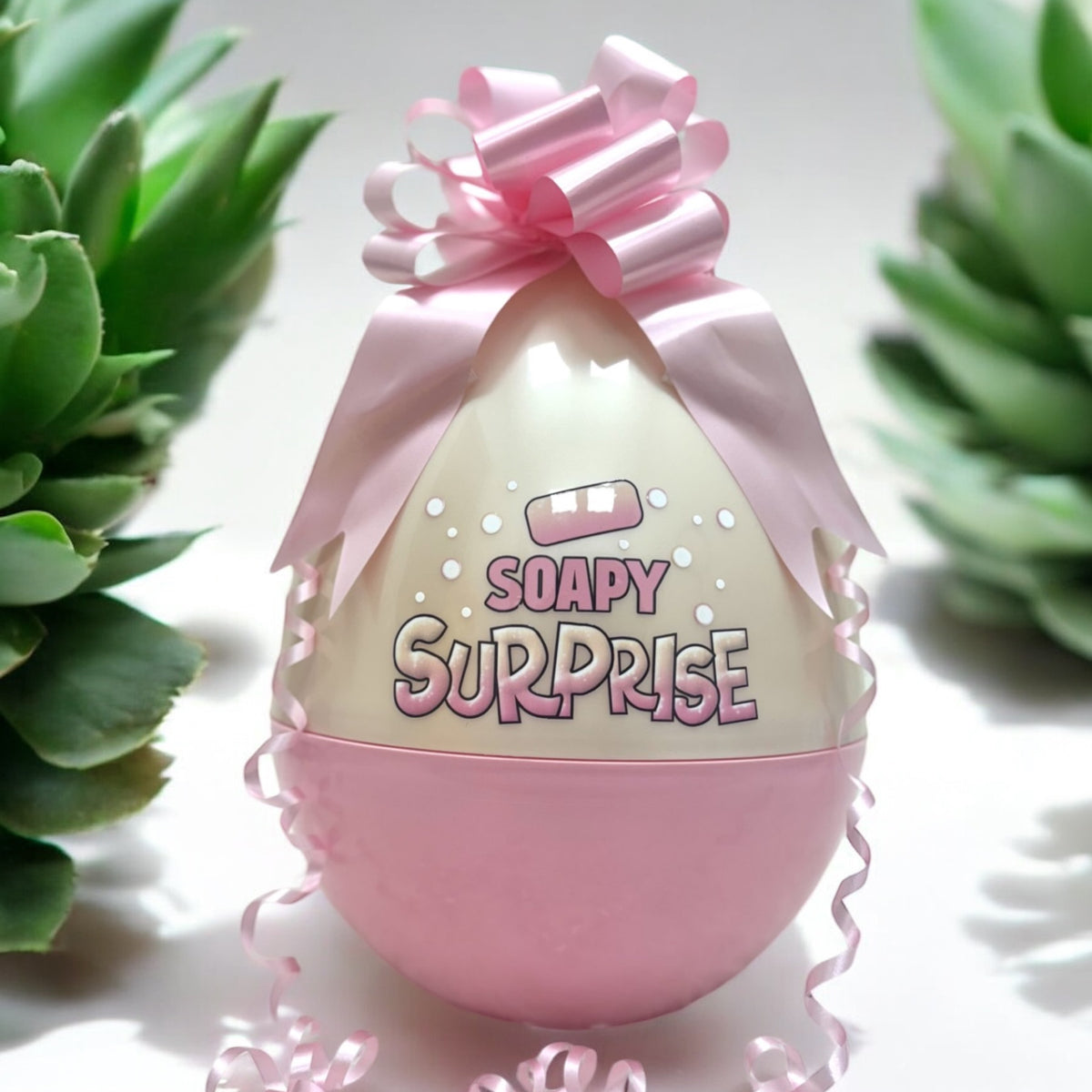 A pink Easter egg with the text 'SOAPY SURPRISE' on it, decorated with a pink bow and placed on a surface with green plants in the background.